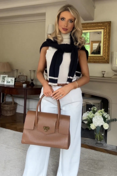 A woman looks in the old money aesthetic style. She is wearing a white knit top, classic white pants with a brown belt and purse. A dark blue knit sweater is tied around her shoulders.